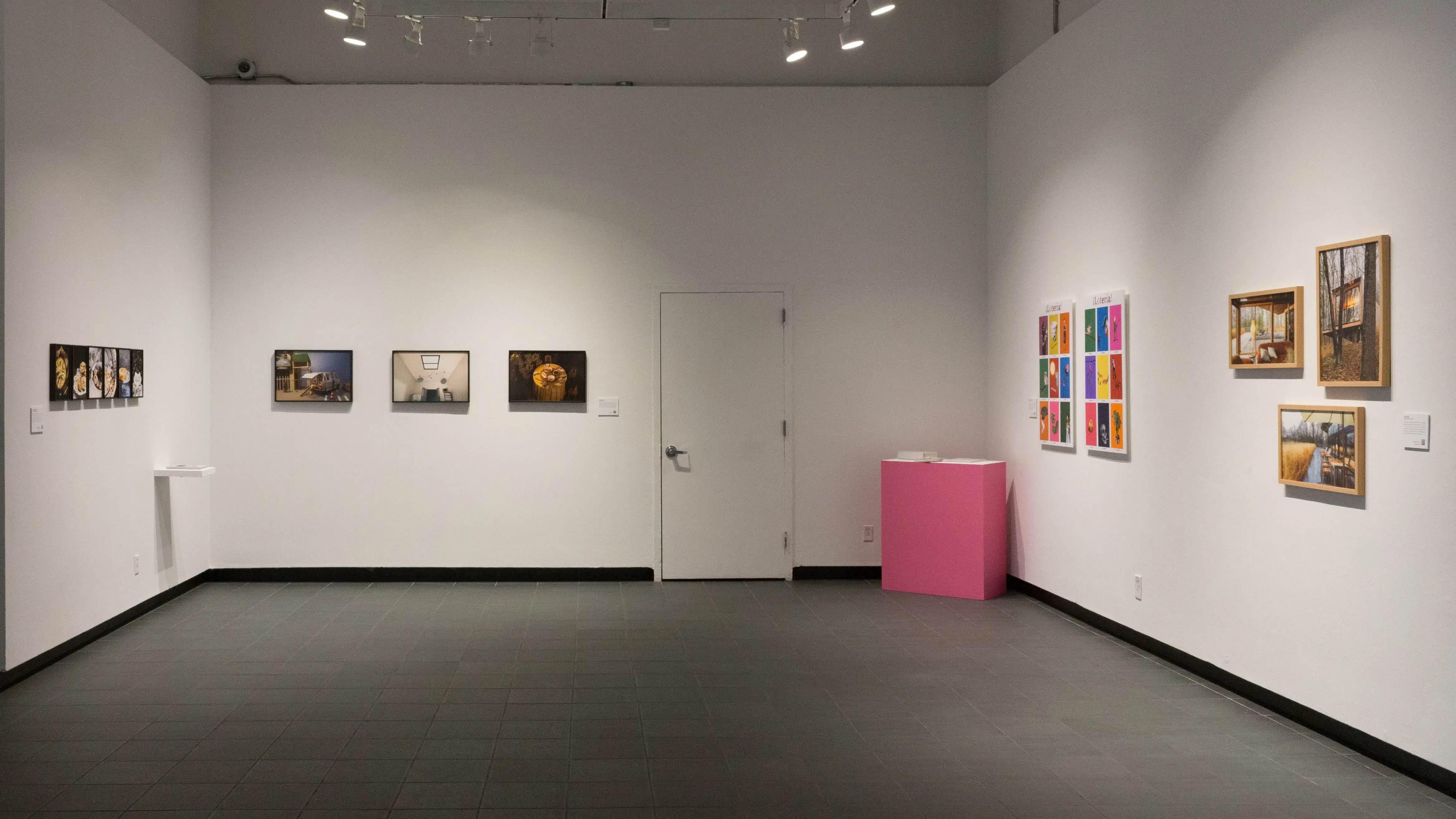 Small framed photos hang in a row on the left wall of the gallery. A few slightly larger framed photos hang in a row to their right, on the center wall. On the right wall, a pink pedestal sits next to two colorful prints and framed photos containing neutral colors hang on its right.