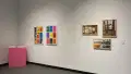 In the corner of the gallery, papers are stacked on a pink pedestal. To its right, two colorful prints with the words “!Lotería!” are hung on the wall. Framed photos containing neutral colors hang further right.
