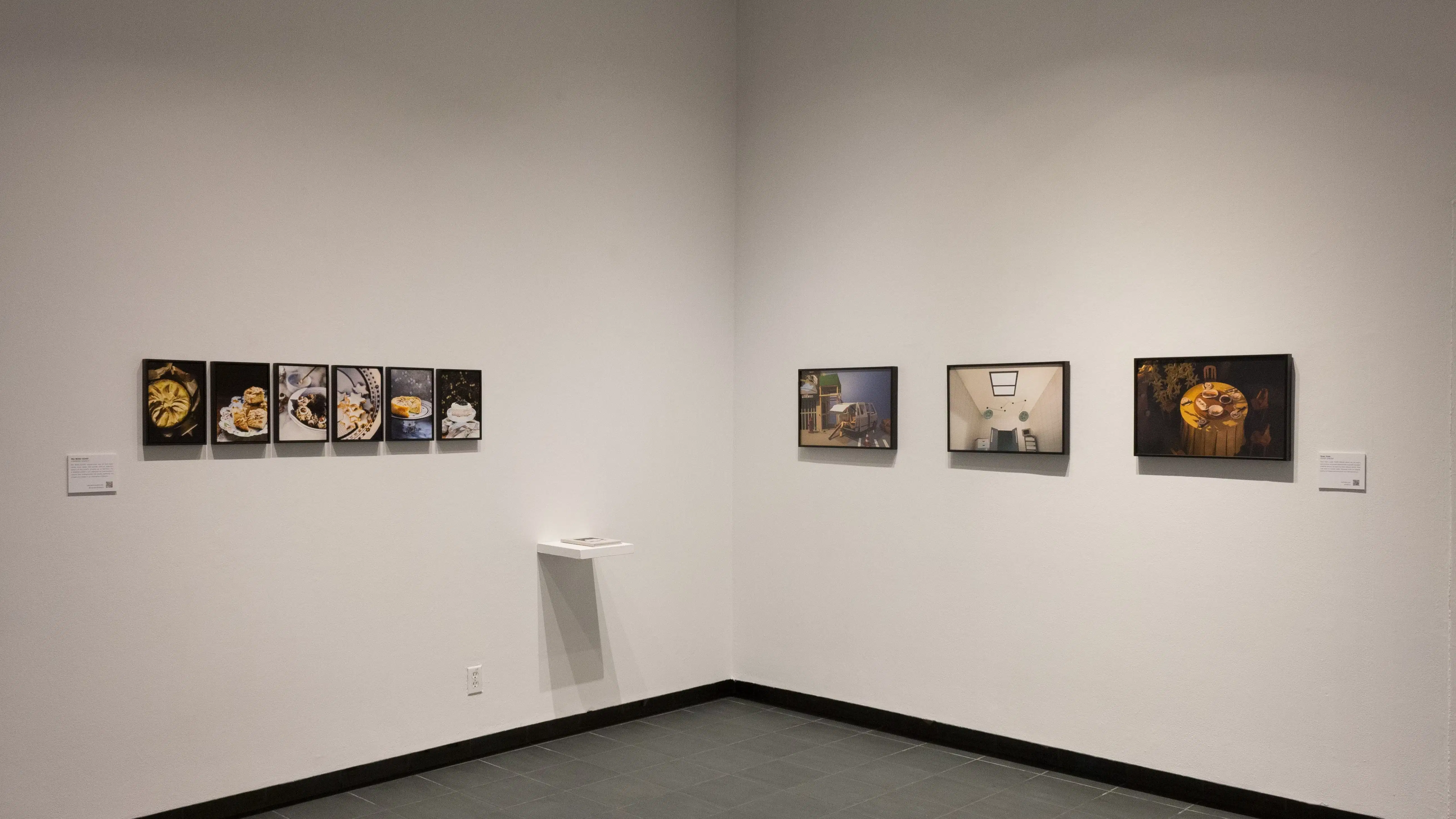 Small framed photos of food hang in a row on the left wall of the gallery. A few slightly larger framed photos hang in a row to their right, on the center wall. 
