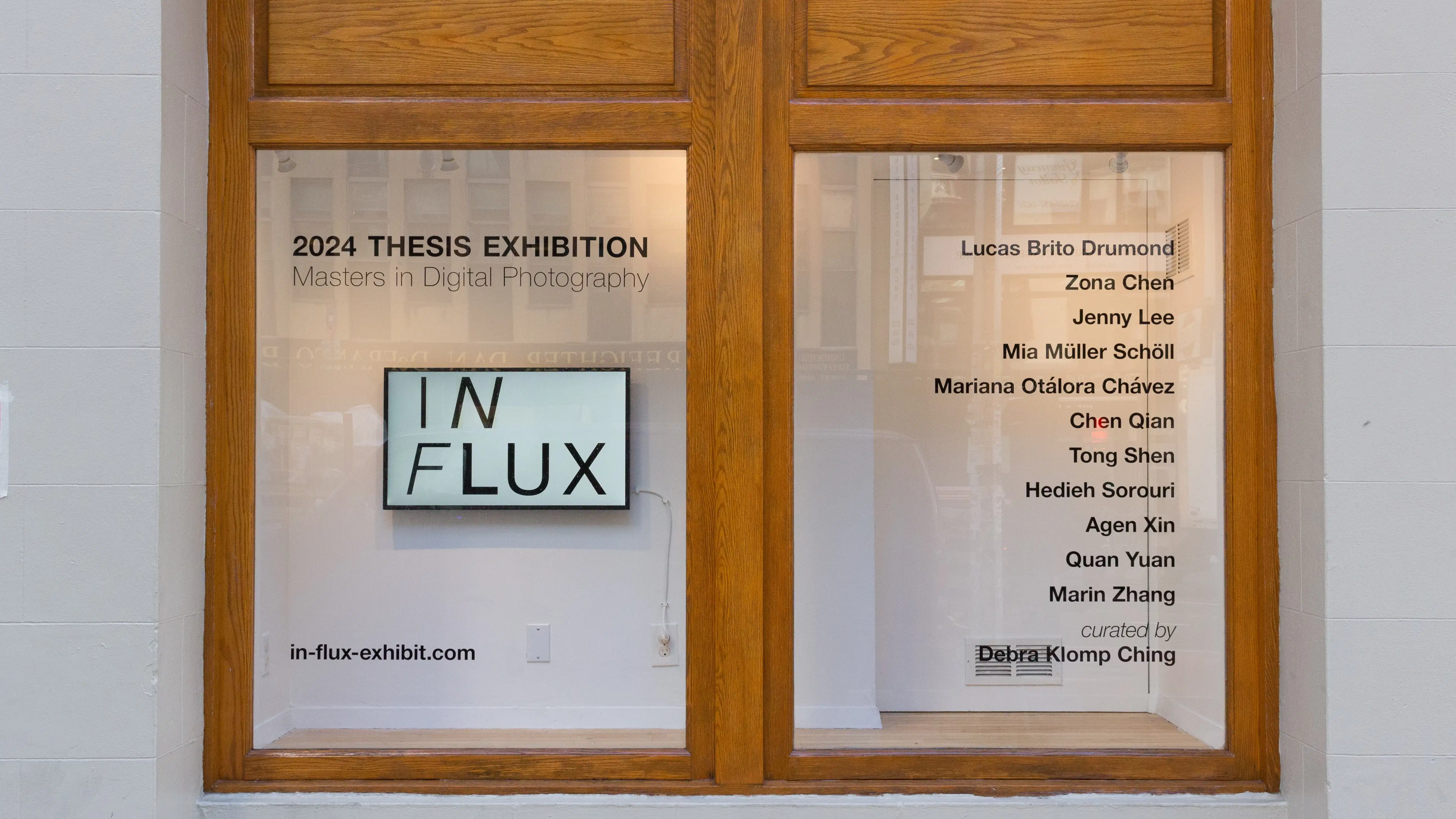 In the display window outside of the SVA Gramercy Gallery, a monitor displays the title of the exhibition “In Flux.” Window vinyl shows the list of participating artists.