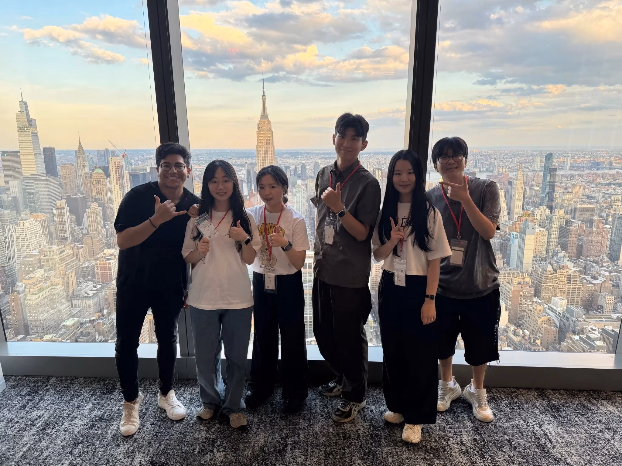 A group of five students and a professional stand in front of a large floor-to-ceiling window overlooking the New York City skyline at sunset. The Empire State Building is centered in the background. The group smiles and poses casually with hand signs and peace gestures, each wearing name badges on red lanyards.