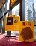 Two yellow boards with orange patterns stand in a sunlit atrium with large windows showing a city skyscraper. The display on the left has a black digital monitor with the text "FALAK KHOJA" and some information underneath. A vertical board on the right has the text "TLK X SVA" and "ARTWORK BY ELYZA NACHIMSON" above a dark brown, laser-cut design featuring a stylized lion on a mock manhole cover.