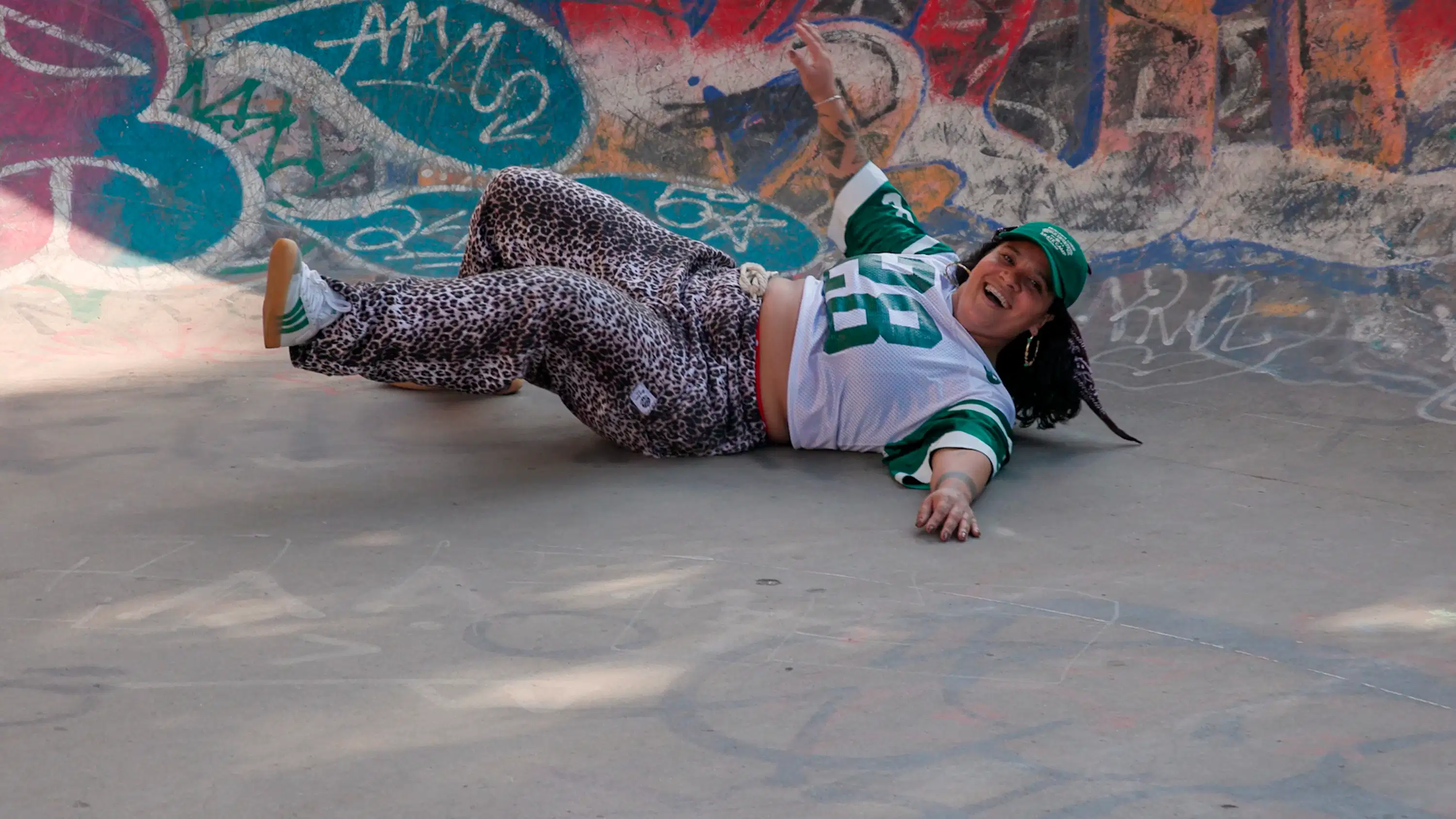 Color still from FREE SKATE (dir. Lia-Alexi Manfredi, 2025). A woman in leopard-print pants and a green and white sports jersey lies in a graffiti-covered skate bowl, laughing after a wall with one arm raised.