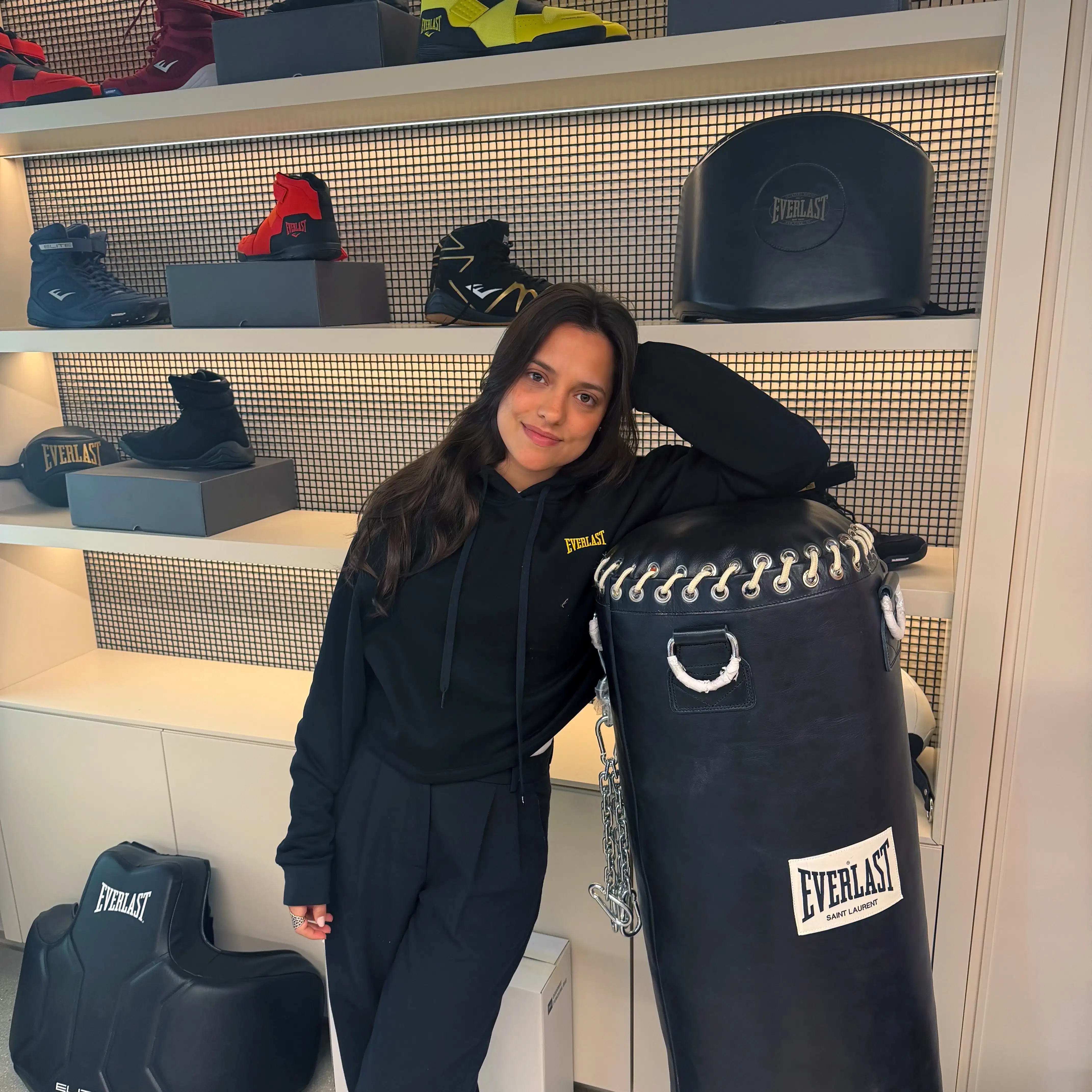 A brunette woman in black workout sweatshirt and pants is standing by an Everlast boxing display room showing a punching bag, shoes, and equipment on shelves. She leans against an Everlast-branded punching bag., 