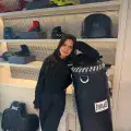 A brunette woman in black workout sweatshirt and pants is standing by an Everlast boxing display room showing a punching bag, shoes, and equipment on shelves. She leans against an Everlast-branded punching bag., 