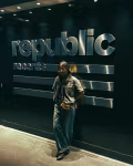 A student stands in a dimly lit hallway leaning against a wall with the large metallic “Republic Records” logo, wearing glasses, a short-sleeve button-up shirt, wide-leg jeans, and a crossbody bag.