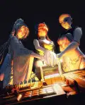 A dramatic, stylized illustration of three women—Yuki, Lena, and Lilian—standing shoulder to shoulder in a dimly lit, high-tech setting, have their hands placed firmly on a large, glowing control lever as if preparing to activate a powerful mechanism. 