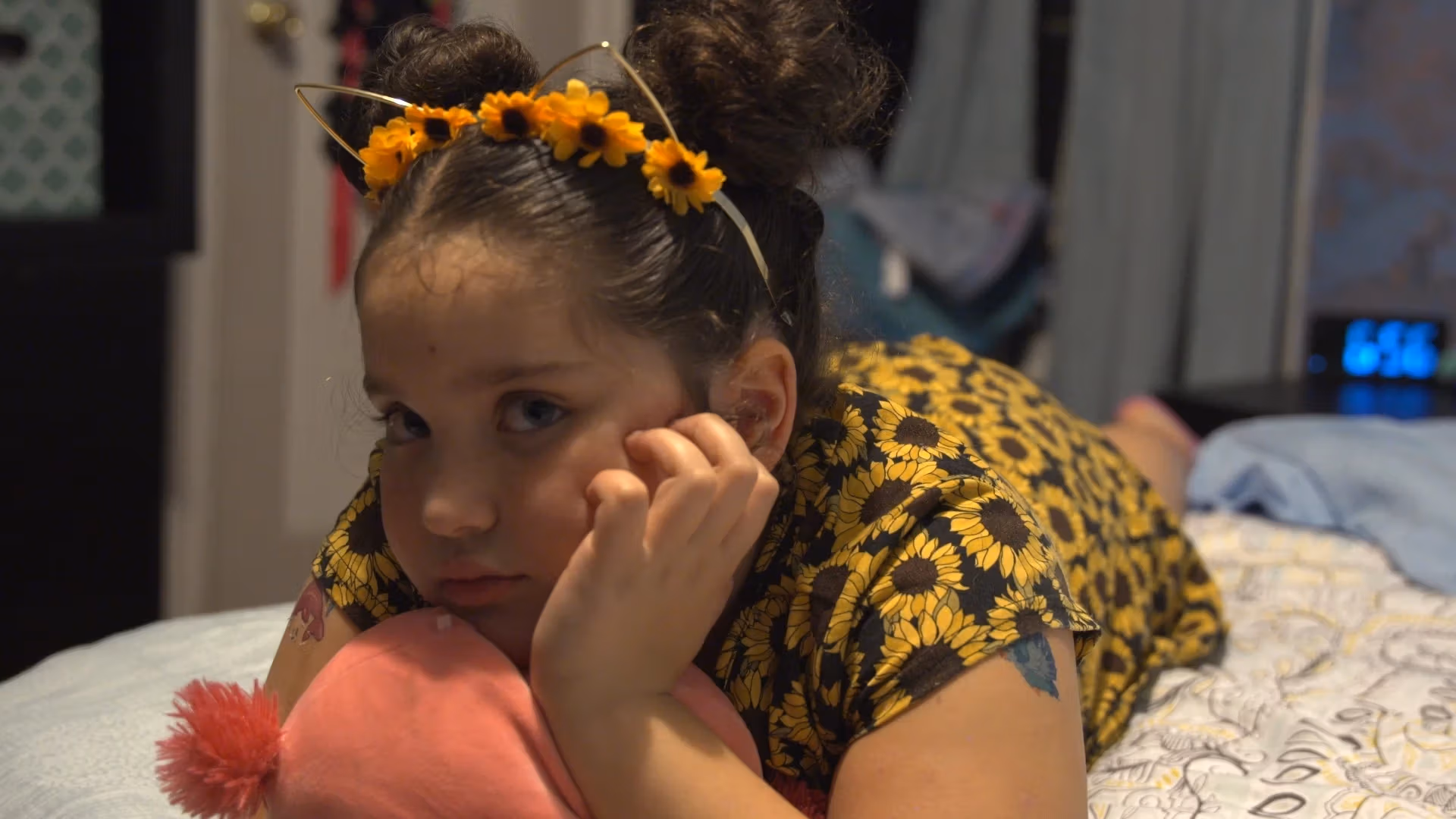 little girl in sunflower dress and sunflower headband lies in her bed with her hand under her chin and a serious expression