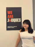 A young woman stands indoors next to a wall poster that reads “WE ARE JACQUES” in bold red and white text, with the word “Havas” incorporated into the design. Below it, smaller text says “CREATIVITY HEALS EVERYTHING.” She is looking to the side with a slight smile, wearing a white off-the-shoulder top against a plain light-colored wall.