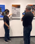 A young woman with a ponytail points to a lunar-themed artwork displayed on a gallery wall while a man films her with a smartphone mounted on a stabilizer. She is wearing dark jeans and a black shirt, and the artwork she gestures toward shows a rover on a textured moon surface with an orange planet in the background. Additional framed artworks hang on either side of the piece.