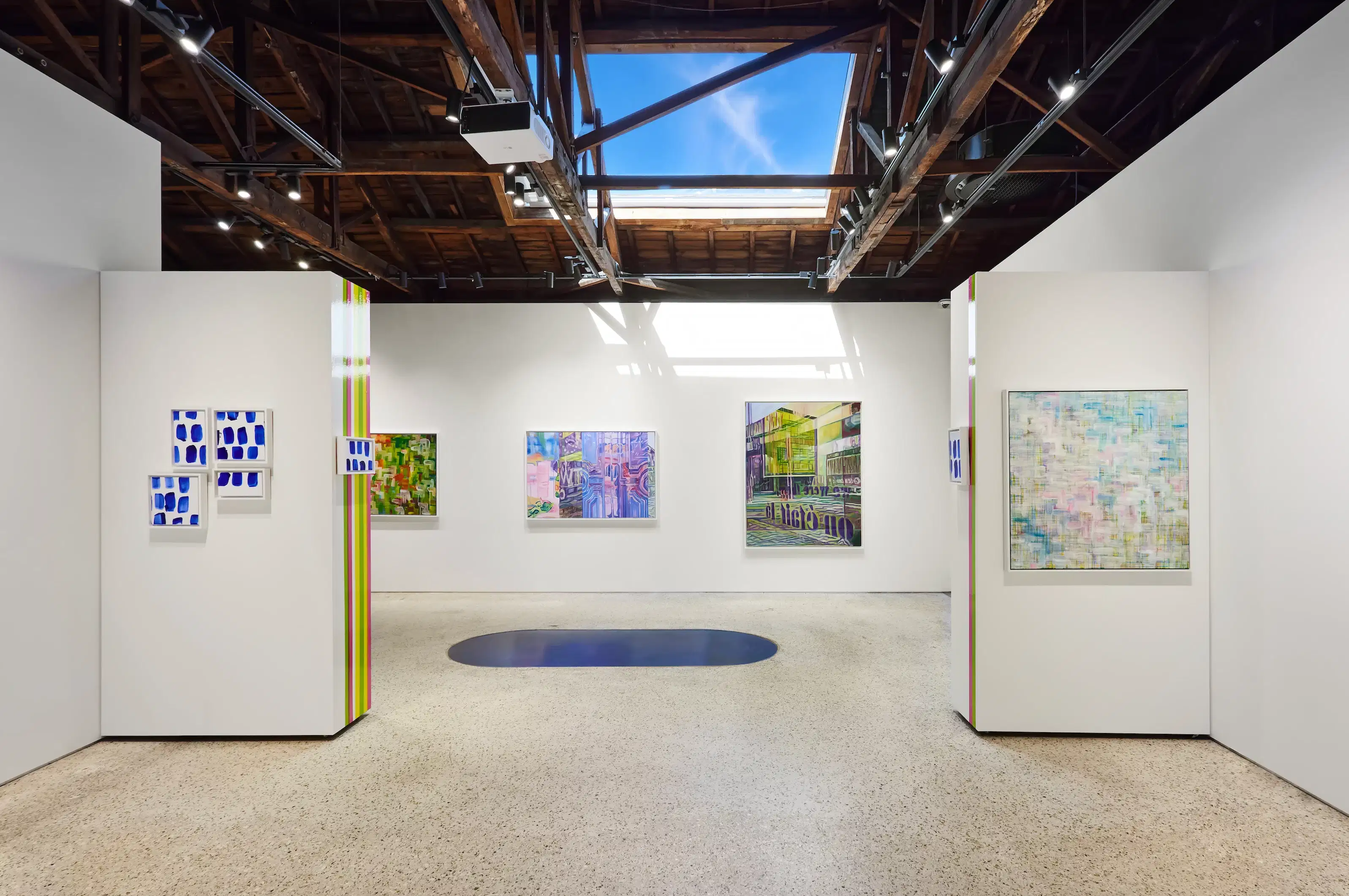 Contemporary gallery space showcasing abstract and geometric artworks in vivid, layered colors. Paintings are hung on crisp white walls beneath a wooden beam ceiling with a skylight that casts natural light across the space