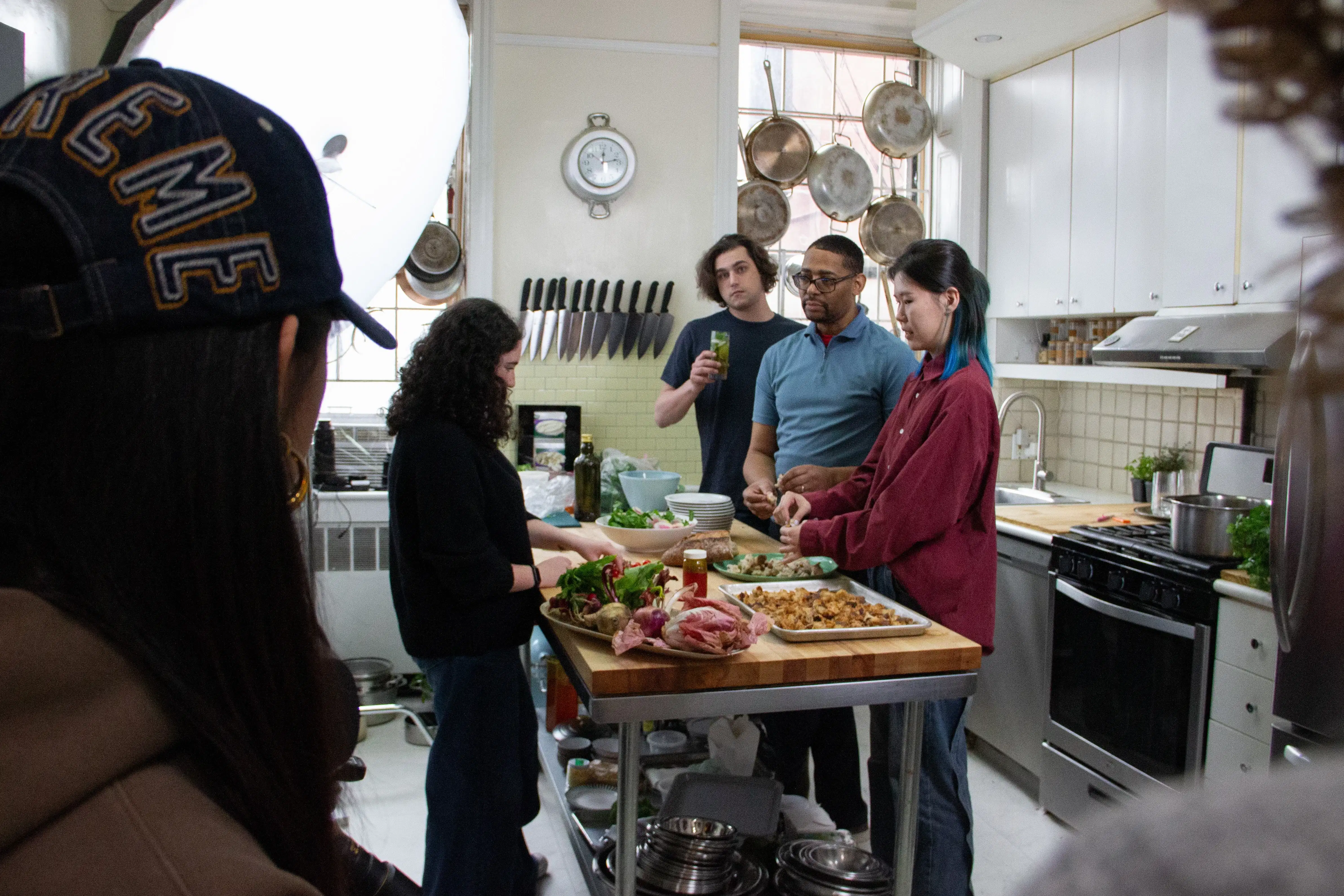 A behind-the-scenes shot shows students and staff standing on the right side of a kitchen table next to a large studio light.