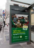 A city bus shelter on a sidewalk displays a green WildAid food-waste poster showing a group of people cooking together in a home kitchen, with bold yellow and white text that reads “Yesterday’s take-out becomes tonight’s fried rice”, with “Make tasty meals with food scraps” below it and “Great job!” outlined in a white shape in the middle right of the poster.