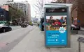 A wide street scene in the city showing a bus shelter that features a blue WildAid advertisement with a group of people standing behind a table full of fresh produce, alongside large white text that reads “Take your food from expiring to inspiring”, with “Make tasty meals with food scraps” below that and “Nice work!,” in a white star in the middle right of the poster. 