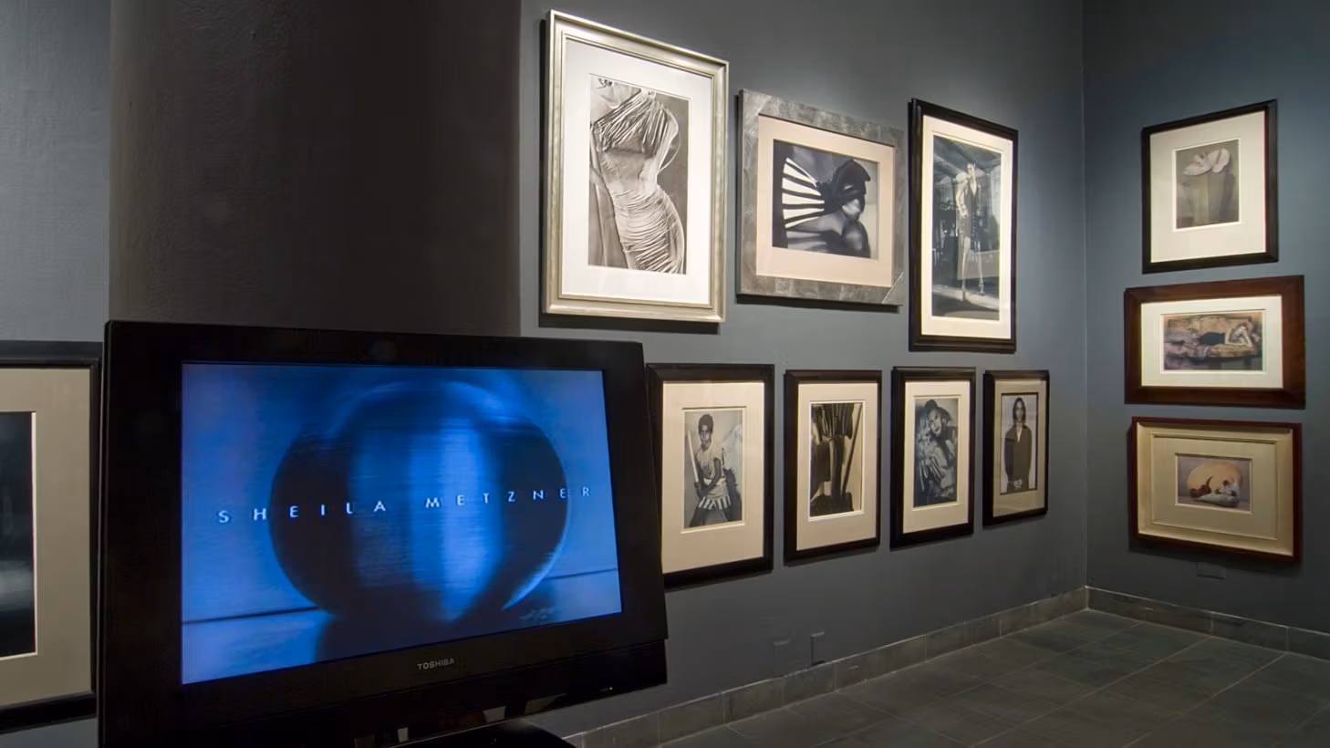 Photograph of wall at an angle, showing seven framed photographs in grayscale and sepia-tone. On the far left, displayed on a monitor, is an abstract image of a metallic orb with "Sheila Metzner" superimposed on it.