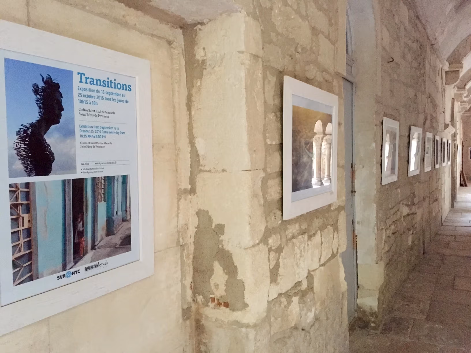 At the entrance of the Romanesque cloister of Saint-Paul de Mausole, several works hang in white frames on a stone wall. The first framed piece is a poster for the exhibition, which is titled "Transitions."