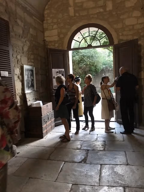 People gather at the entrance of the Romanesque cloister of Saint-Paul de Mausole. The floors and walls are made of stone, and natural light pours through the doorway.