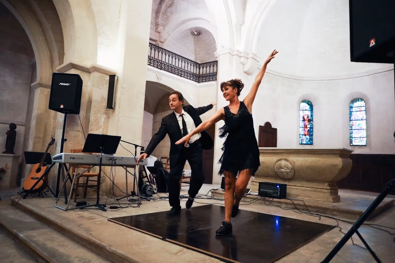 Two performers tapdance on a stage in front of a large dome. One wears a necktie and blazer, while the other wears a fringed flapper dress.