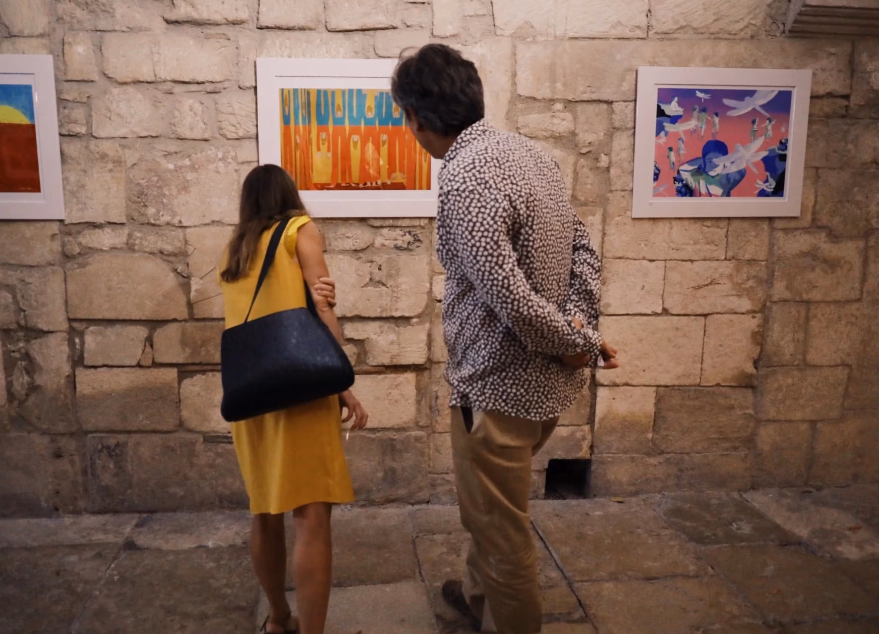 A couple take a close look at a colorful illustration that hangs on the walls. The work is abstract, using yellow shapes on a blue and orange background.