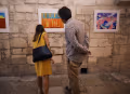 A couple take a close look at a colorful illustration that hangs on the walls. The work is abstract, using yellow shapes on a blue and orange background.