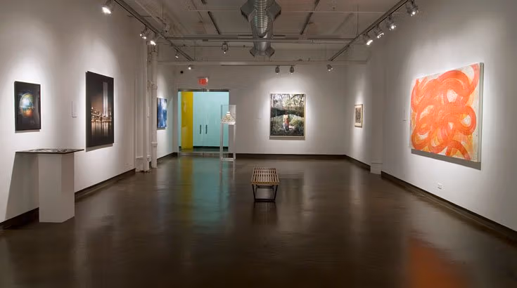 Several different pieces of artwork hanging in a gallery, the one on the right features lots of orange. There is a bench with no back in the center of the room
