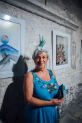 A woman wearing a feathered headband stands among the works on display. She wears blue earrings and a blue dress.