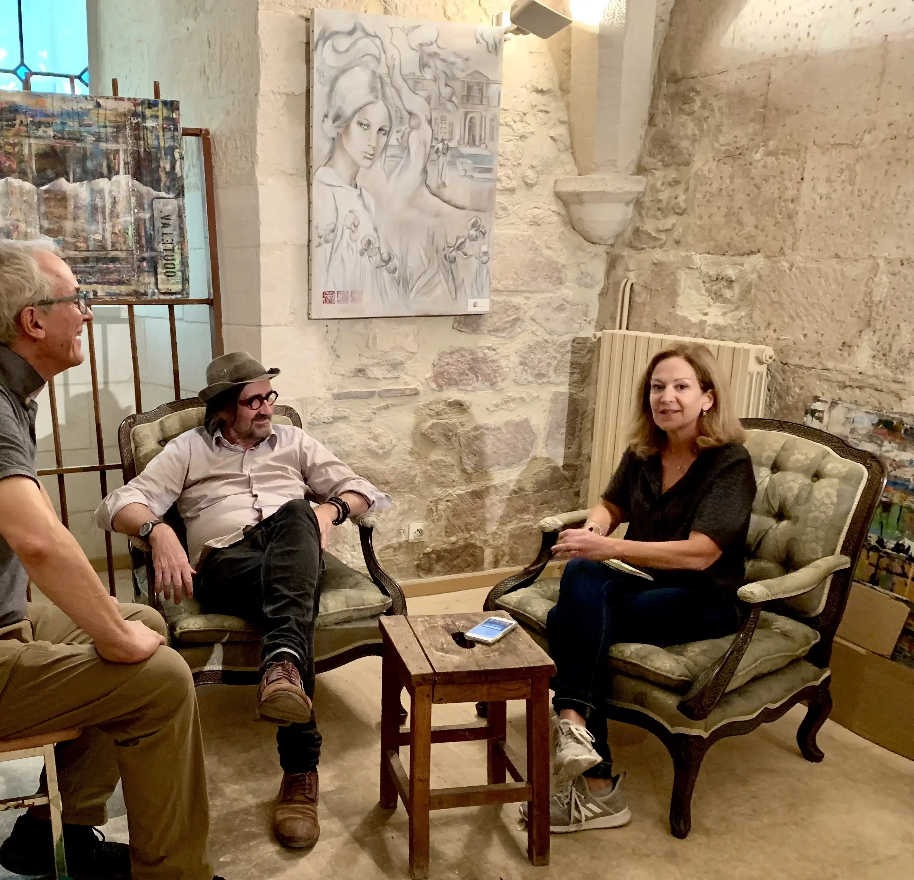 Three people talk and laugh as they sit around a small wooden table. Behind them, two large paintings hang on the walls.