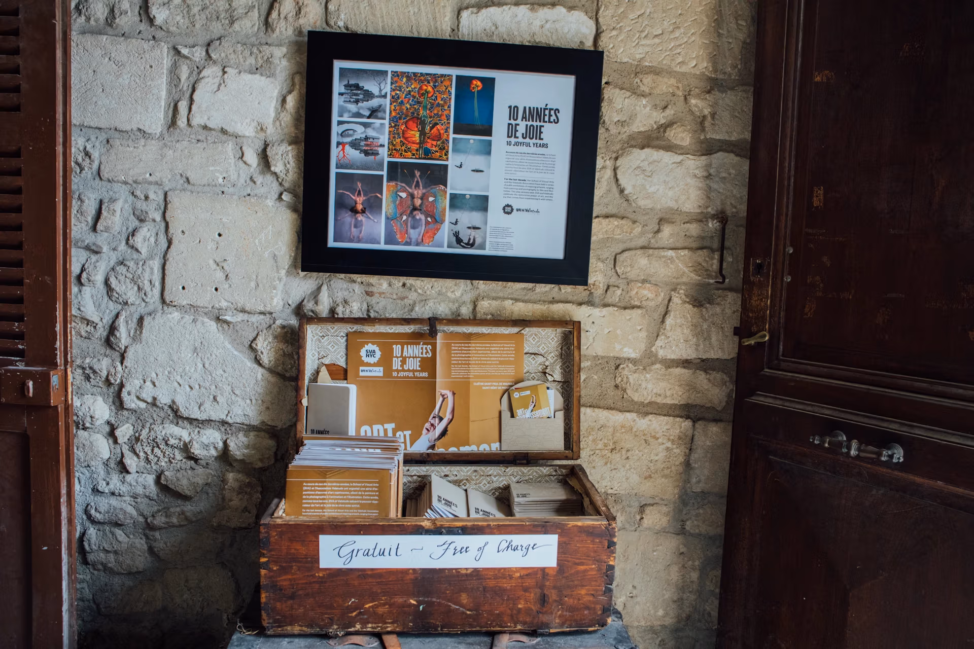 Beneath a frfamed poster for the exhibition, a wooden trunk with a sign that reads "Gratuit - Free of Charge" holds posters and other takeaways.