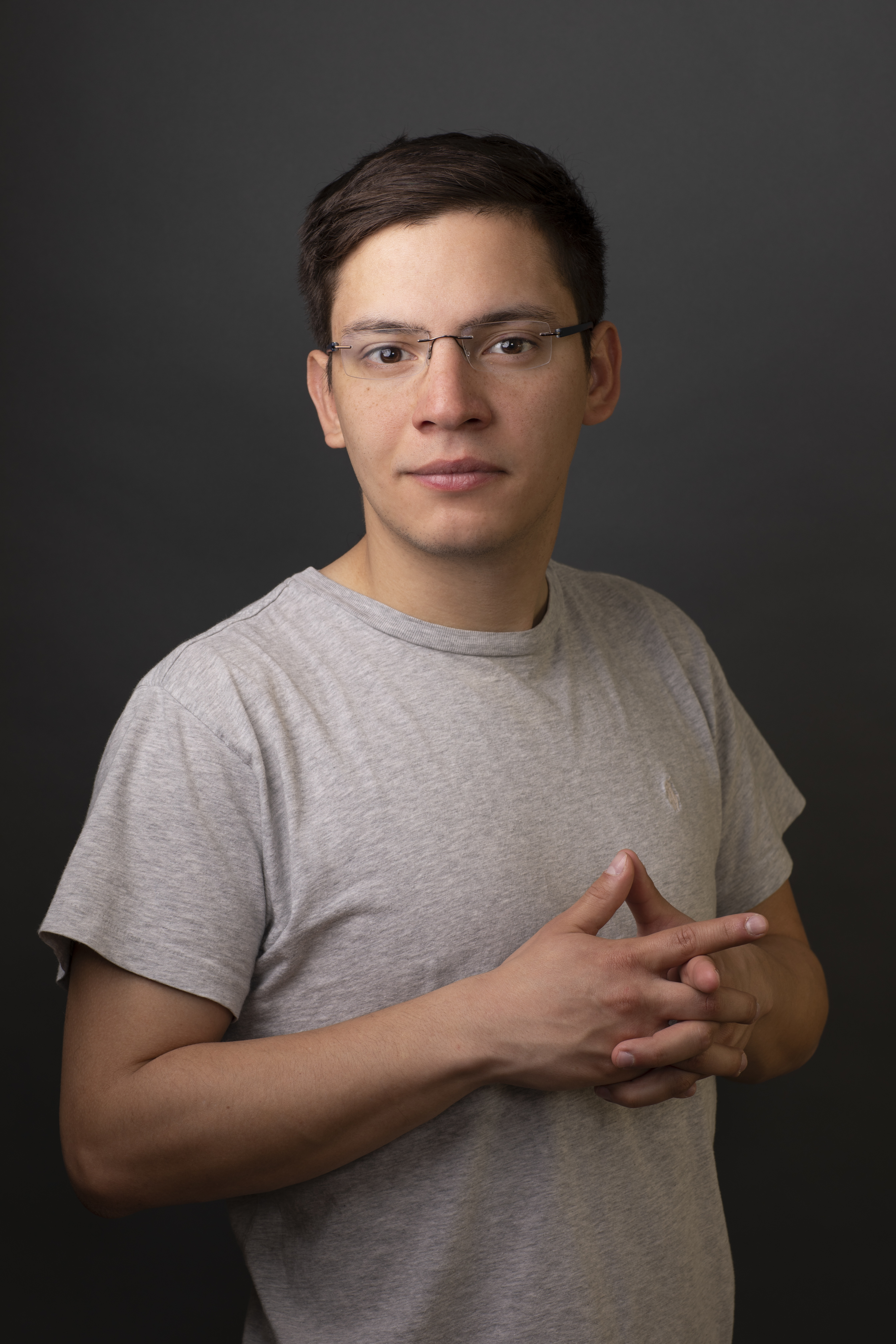 <p "="">A color photograph of a young man standing in front of a monochrome backdrop, with his hands clasped at his chest.