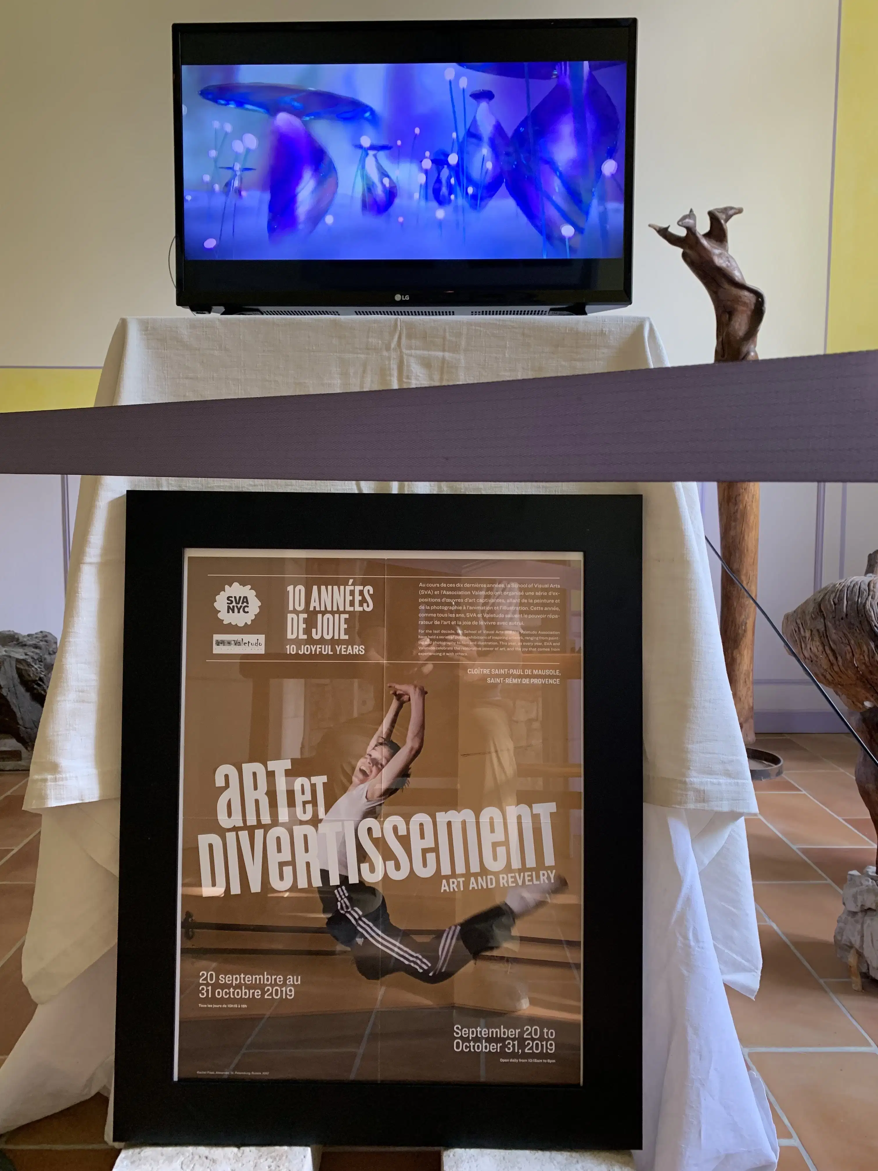 A monitor plays animations, and below the monitor, a framed poster reading "Art et Divertissement" or "Art and Revelry" sits on blocks.