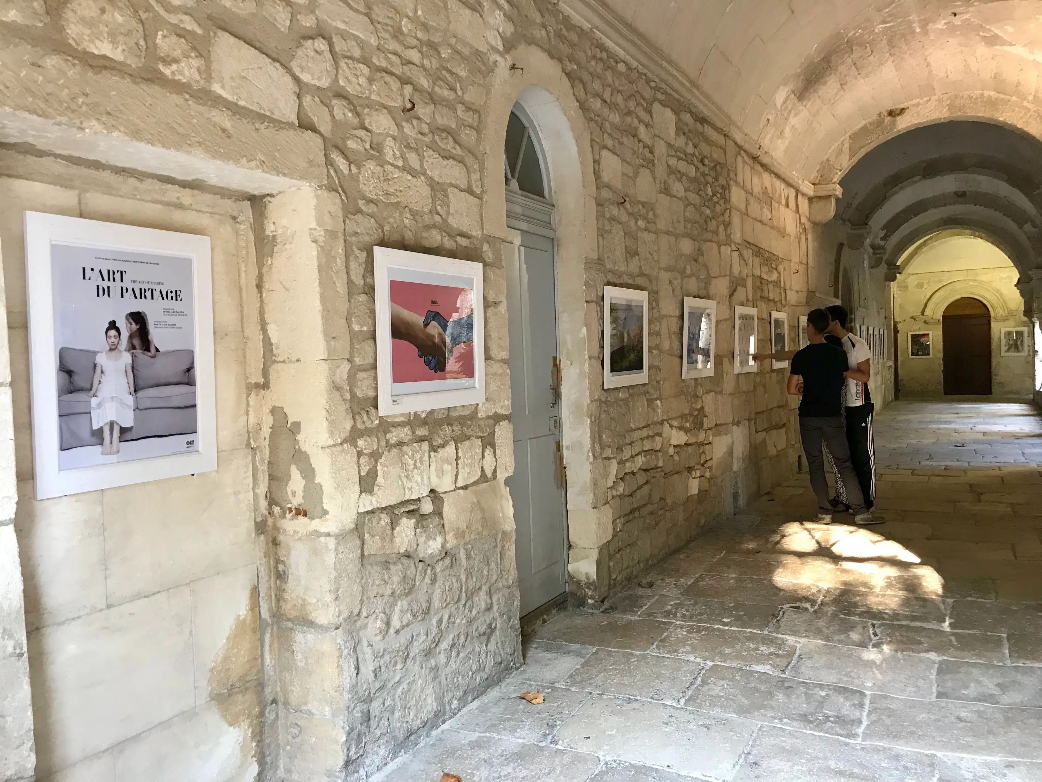 Several framed works hang in a row down a stone wall. The first framed piece is a poster for the exhibition, which is titled "The Art of Sharing"