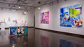On the right is a large blue painting with a young girl looking at viewer, another painting by same artist in pink and purple is to its left, and on far left are multiple shelves and pedestals containing ceramic works. In the center of the space is a large wooden sculpture in a street art style depicting a $20 bill.