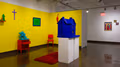 On the left is an inset space, painted yellow, with hyper-pigmented painted objects common to a household including a nightstand, chair, and large dollhouse. On the right are three small, colorful embroidery pieces on a nearby wall.