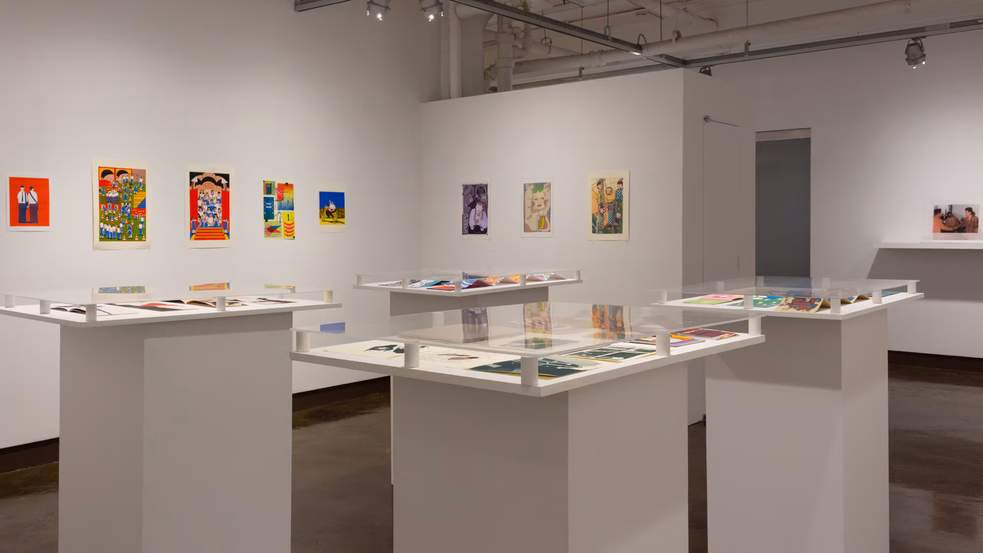 On the inset space to the left are multiple colorful prints in a horizontal line across two adjacent walls. In the center of the space are four white pedestals, each containing multiple printed books, with plexiglass each of the four surfaces.