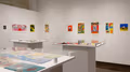 In the corner of an inset space are multiple colorful prints in a horizontal line across two adjacent walls. In the center of the space are three white pedestals, each containing multiple printed books, with plexiglass each of the three surfaces.