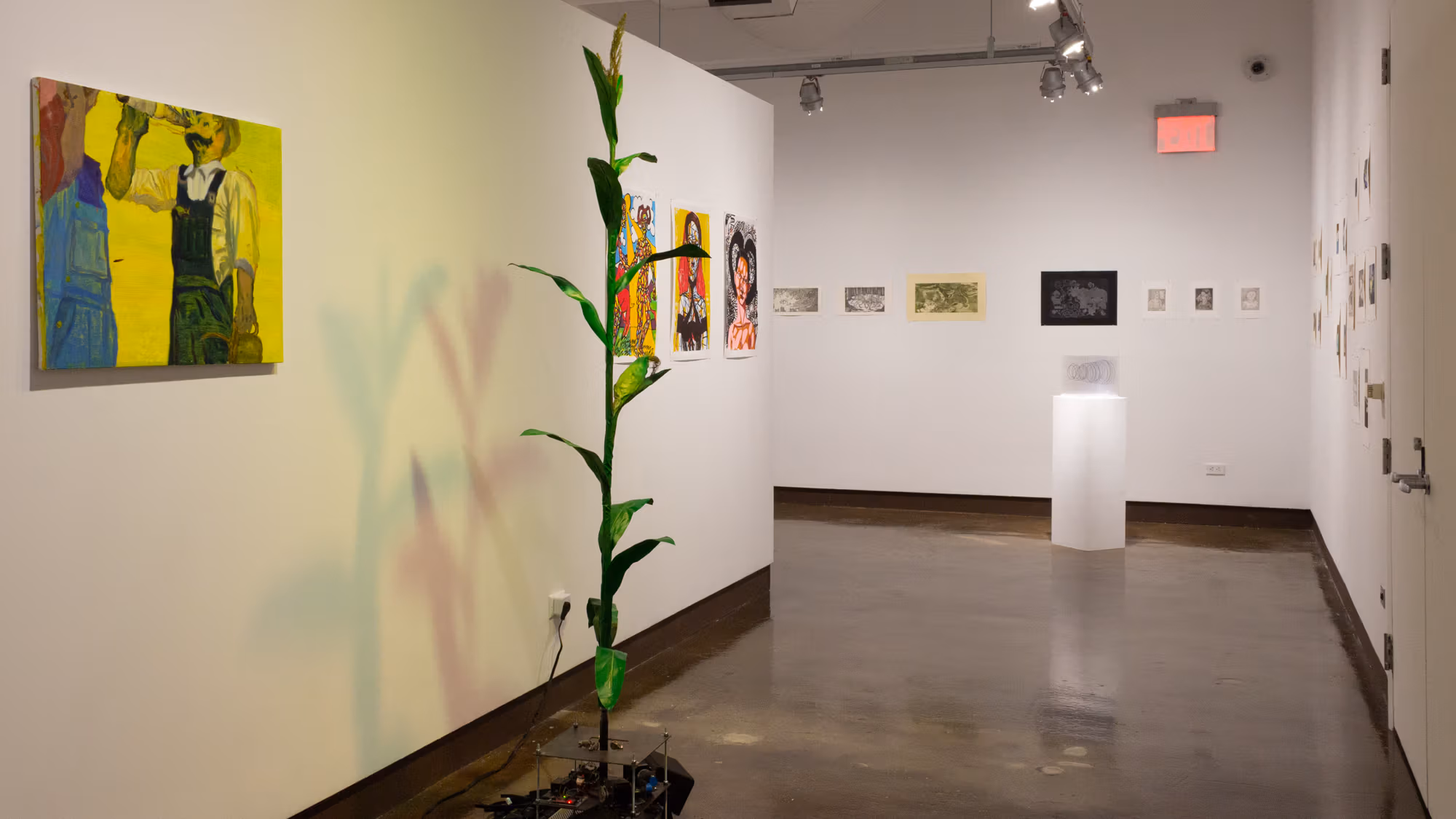 On the left is a yellow-tinted painting of two people drinking from bottles, to its right is a tall plastic cornstalk connected to motors and sensors, and beyond that are many colorful prints hung across the three visible walls.