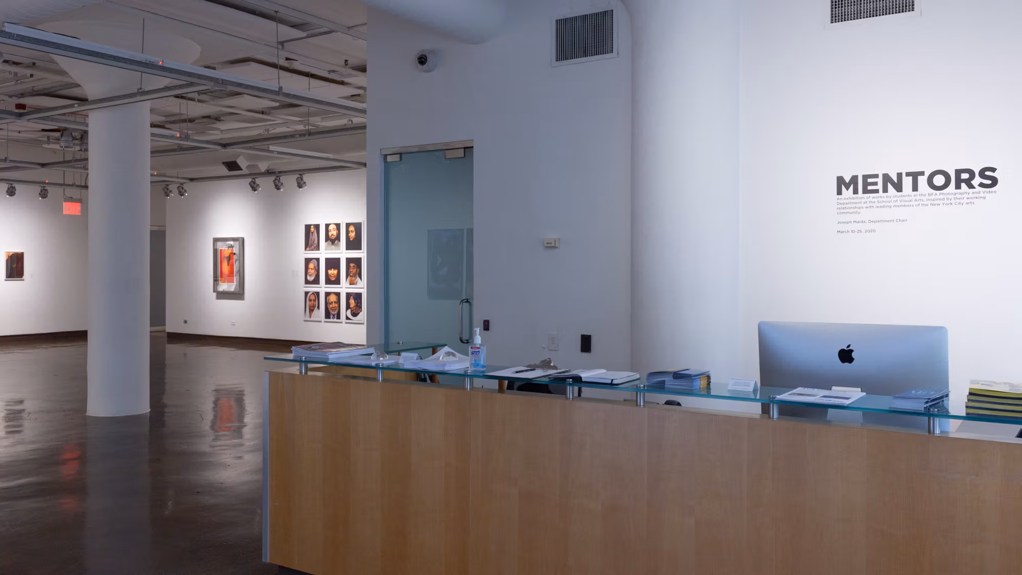 Front desk of SVA Chelsea Gallery, with exhibition title "Mentors" listed in black text behind the desk. On the left is a partial view of artworks in the gallery space.