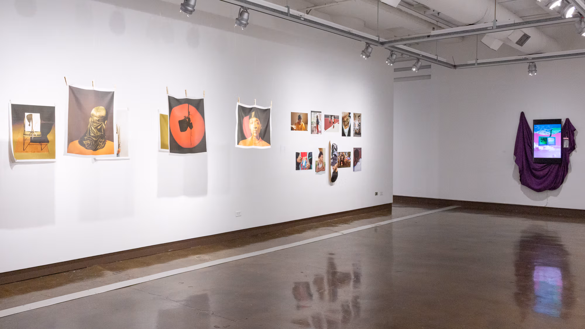 On the left are are six photographs printed on fabric, three of which are suspended and approximately 24 inches from those on the wall. On the right is a six-by-two grid of photographic prints, the bottom center of which is printed on a skateboard, and on the neighboring wall is a monitor presenting a video with maroon drapery behind it.