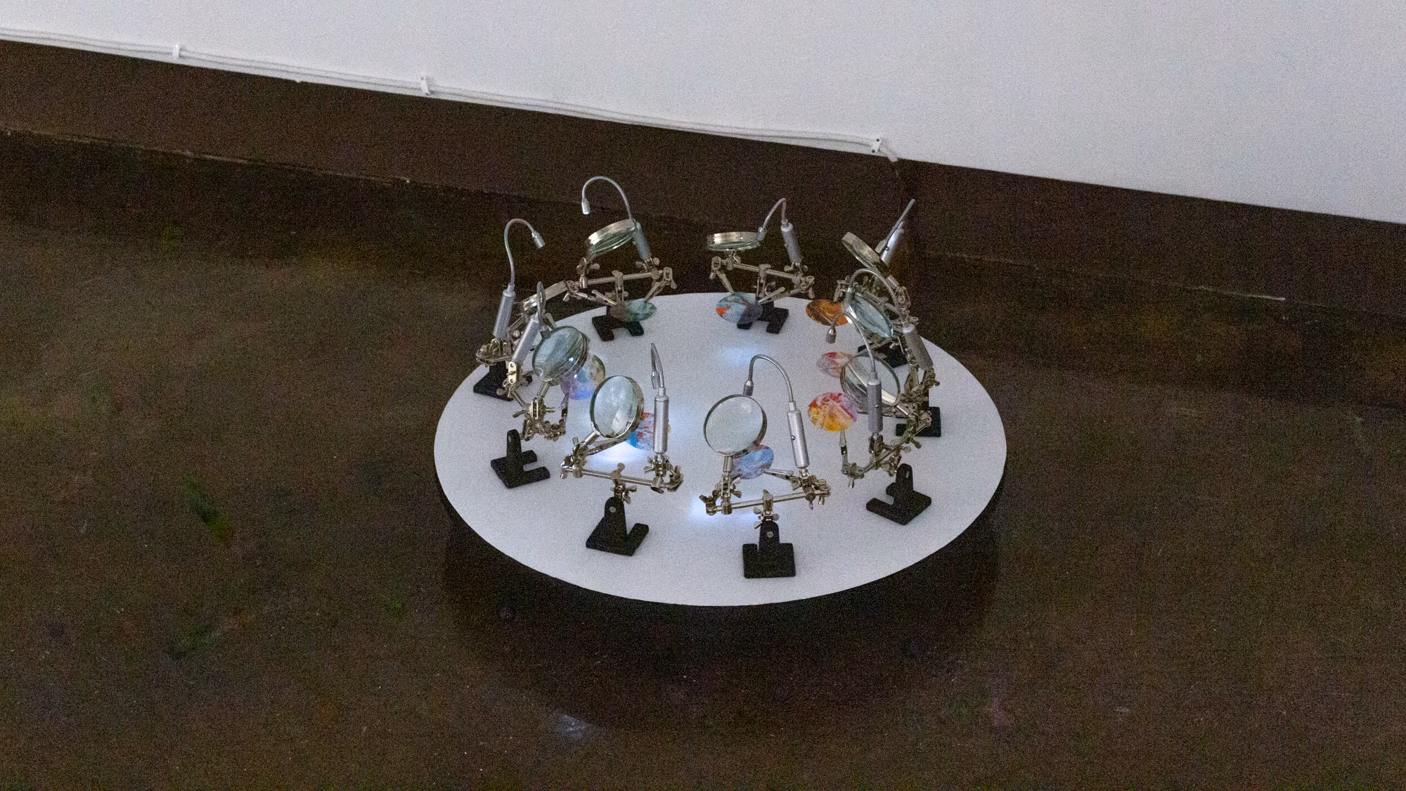 Detail shot of artwork on floor consisting of a "lazy susan" circular disk that spins and a series of illuminated examination tools with magnifying glasses around the perimeter.