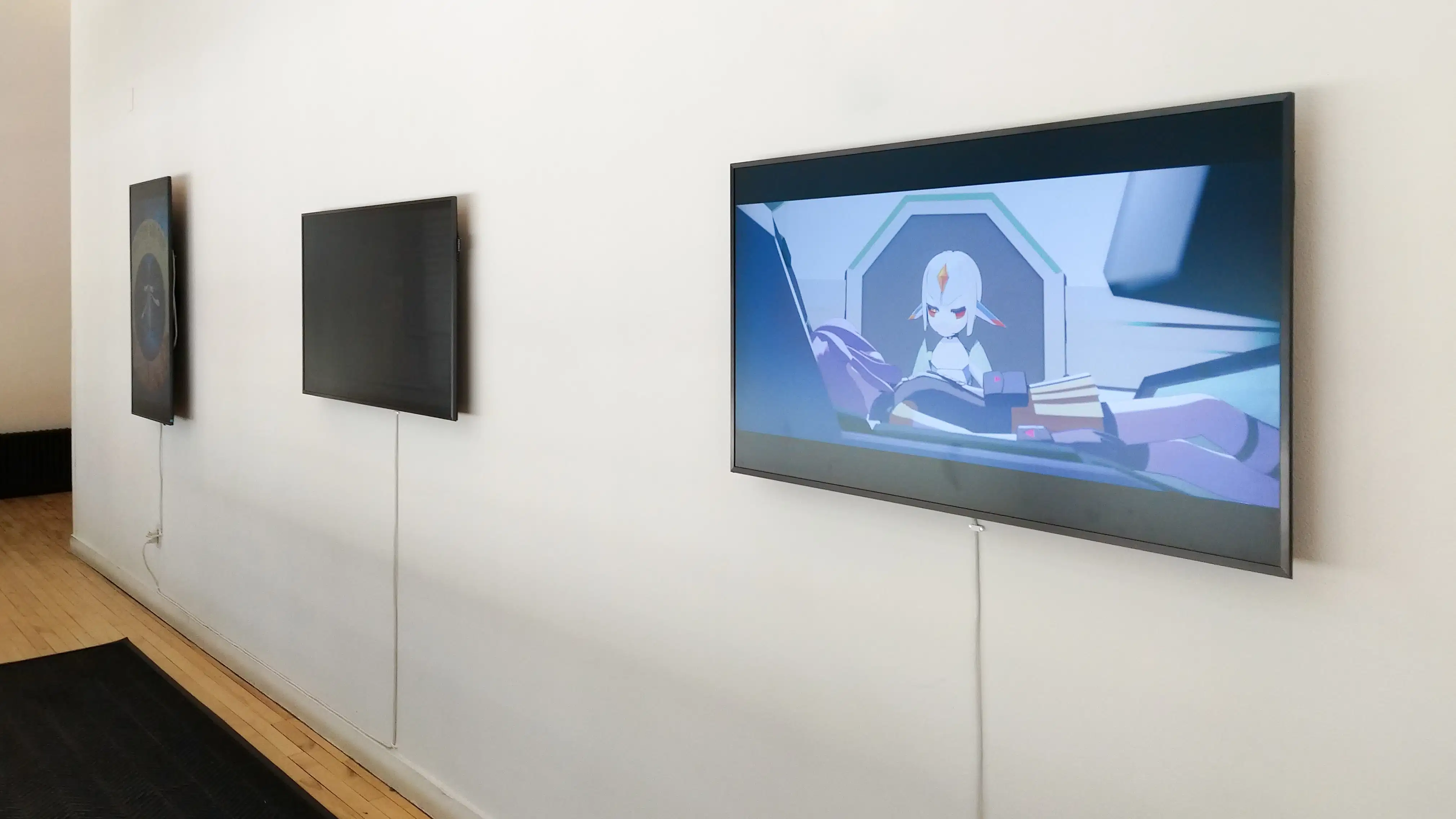 Three video monitors installed on a white wall, the nearest of which plays an anime cartoon.