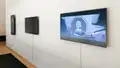 Three video monitors installed on a white wall, the nearest of which plays an anime cartoon.