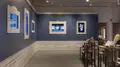 Multiple views of SVA Gramercy Gallery. The lobby is pained red with "Spring Salon" in ornate lettering and the gallery is painted blue. There are monitors in ornate frames playing student work through the gallery. In the center of the lobby and gallery are metal pedestals with student work on them. 