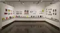 Multiple views of SVA Chelsea Gallery. There is illustrations and paintings hung on the walls. There are multiple shelves throughout the gallery with books places on them.