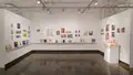 Multiple views of SVA Chelsea Gallery. There is illustrations and paintings hung on the walls. There are multiple shelves throughout the gallery with books places on them.