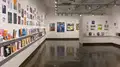 Multiple views of SVA Chelsea Gallery. There is illustrations and paintings hung on the walls. There are multiple shelves throughout the gallery with books places on them.