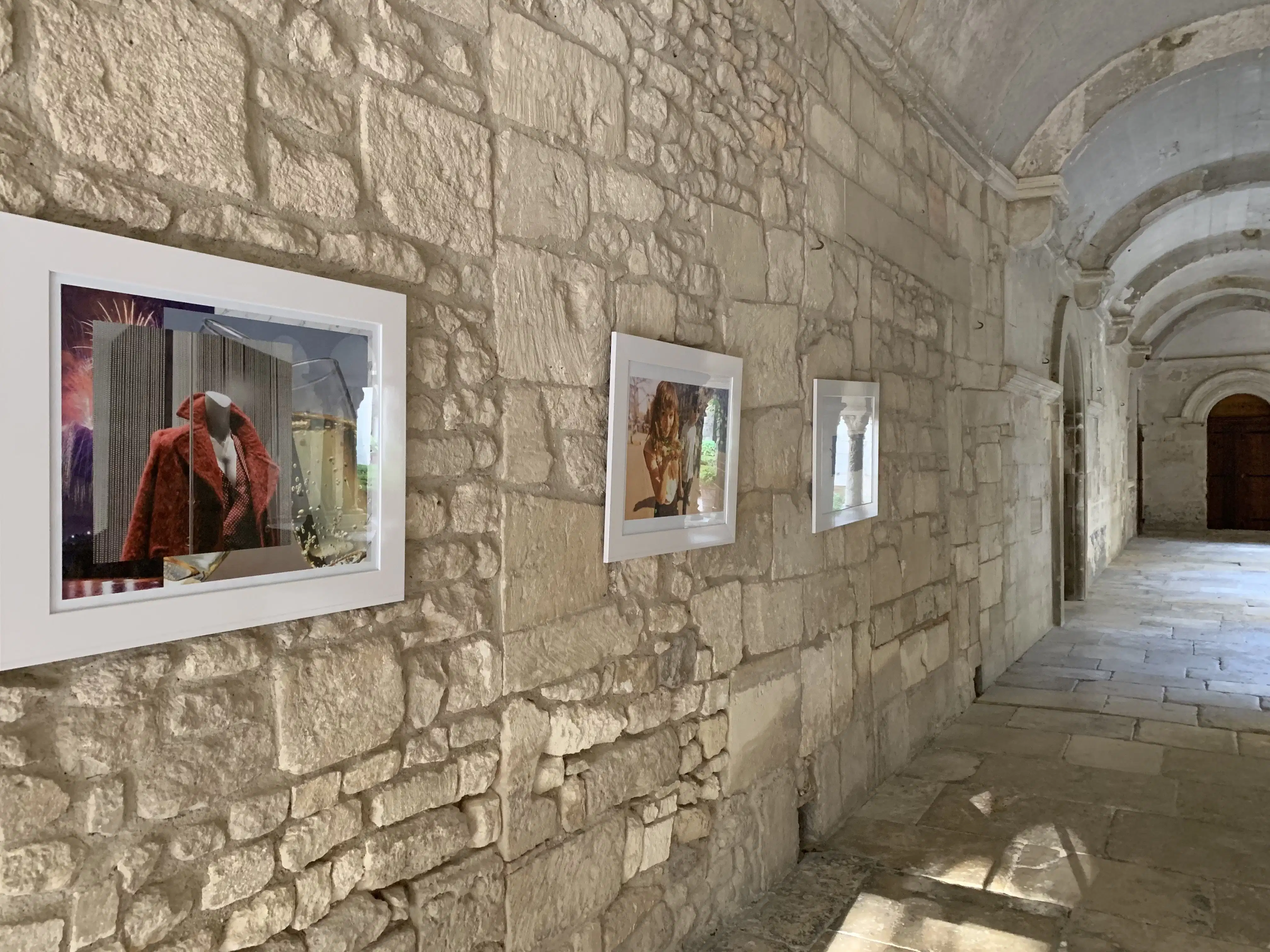 Framed photographs and collages hang in the arched stone hallway. The closest work is a photo of a mannequin, stacked on top of photos of fireworks and champagne glasses.