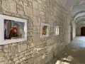 Framed photographs and collages hang in the arched stone hallway. The closest work is a photo of a mannequin, stacked on top of photos of fireworks and champagne glasses.