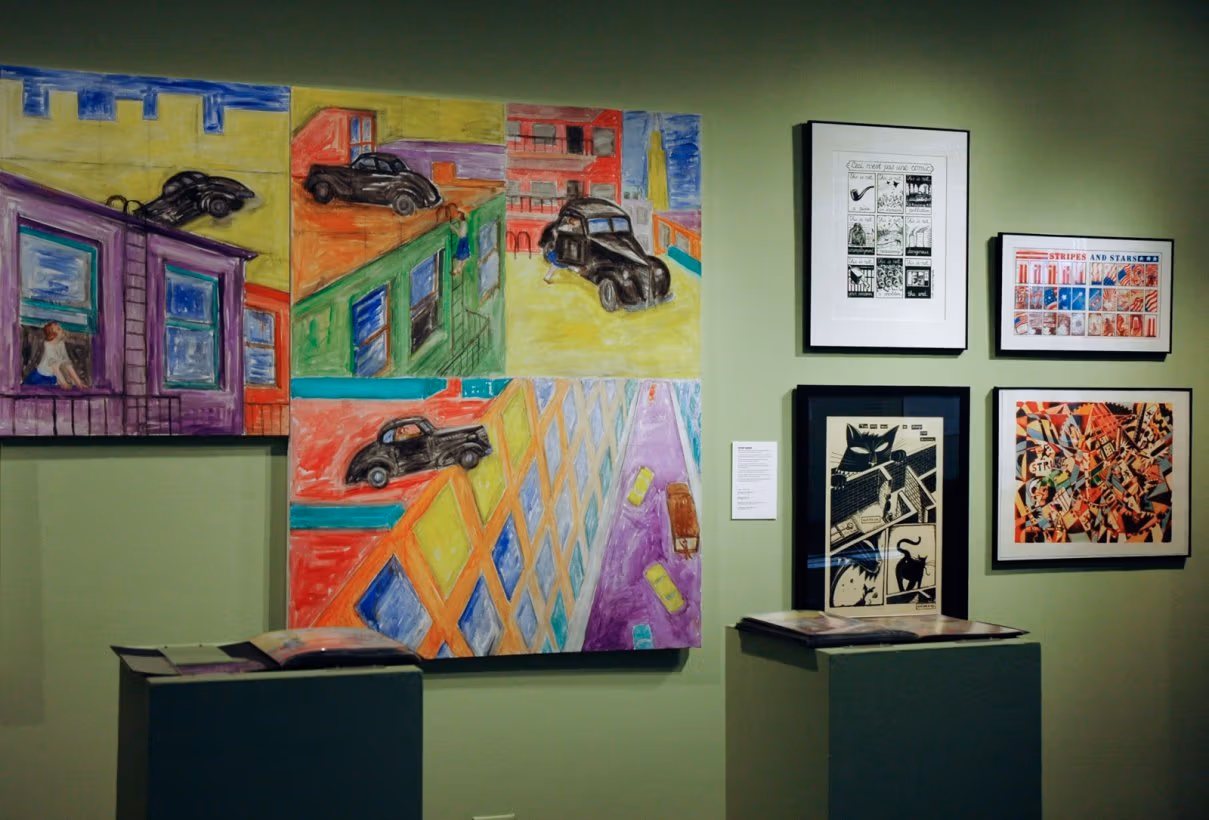 Photo of illustrations hung on green wall, with very colorful ones showing a car on top of buildings to the left and on the right are four more subdued pieces in frames. In front of these are two pedestals containing books.