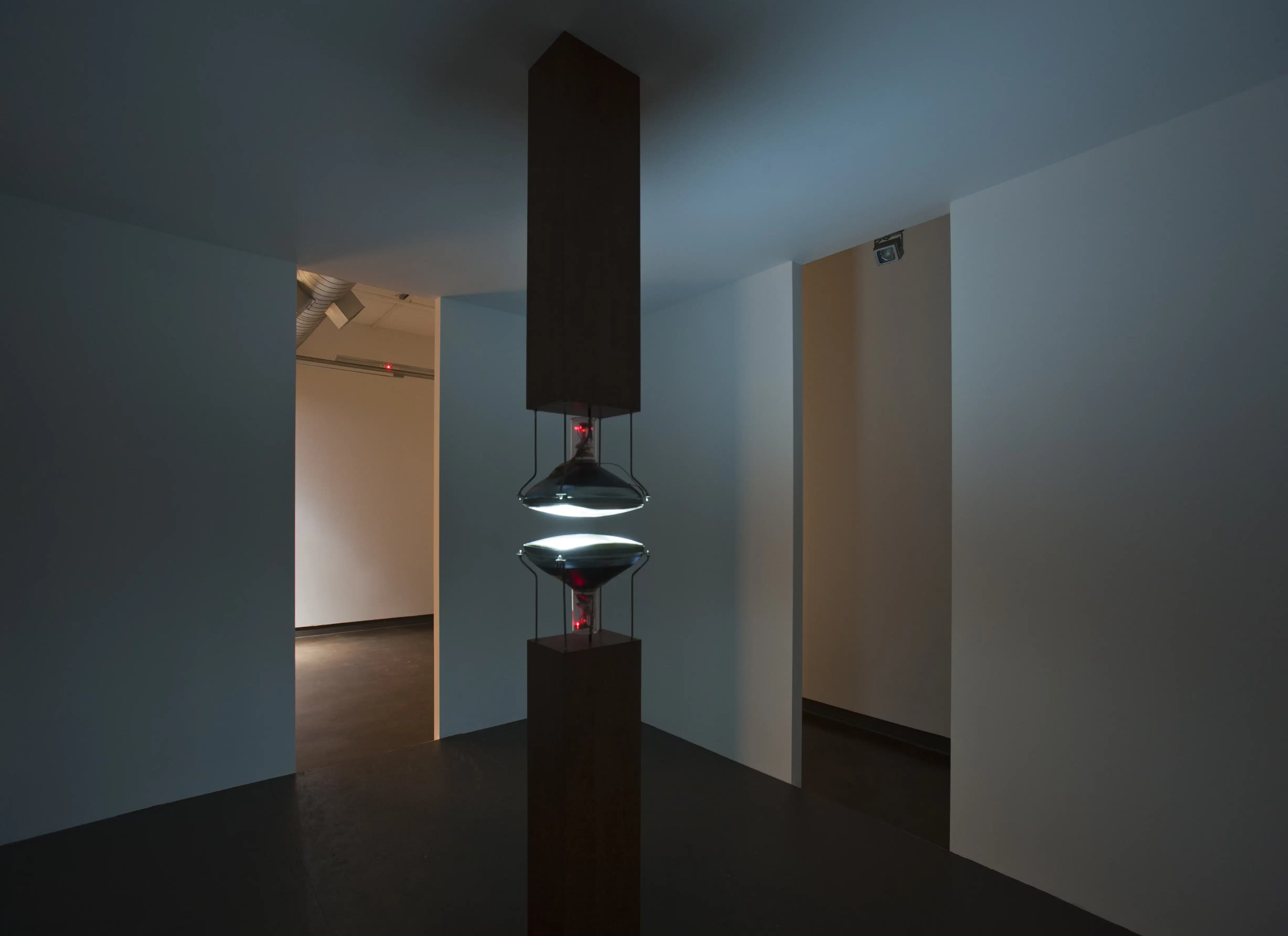 In a white room with two rectangle doors, the center of the room has two lights directly below and above each other on rectangle or bases.