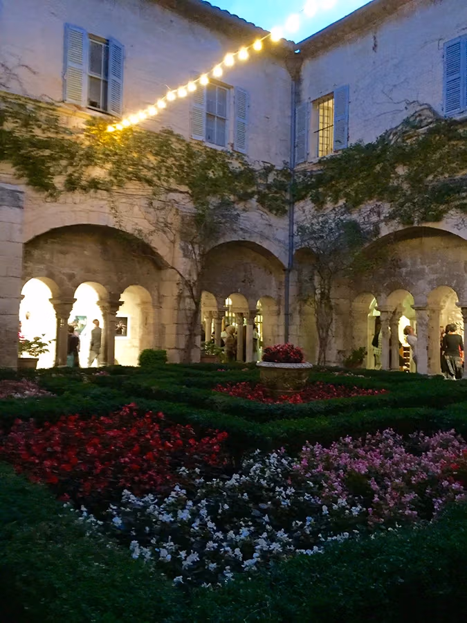 From the cloister garden, the exhibition's works and visitors can be seen through the building's archways. The garden is filled with green hedges and red, white, and pink flowers.